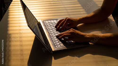 A woman is typing on a laptop keyboard on a white table. The shadow from the blinds falls on the desktop.