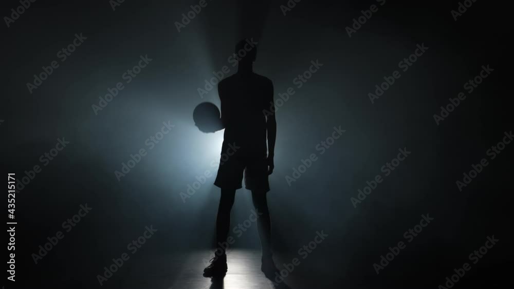 Silhouette of a young guy basketball player tossing the ball up. Tall young sportsman in sports uniform posing on a dark studio background with smoke and backlight. Slow motion ready 59.94fps. Silhouette of a young guy basketball player tossing the ball up. Tall young sportsman in sports uniform posing on a dark studio background with smoke and backlight. Slow motion ready 59.94fps.