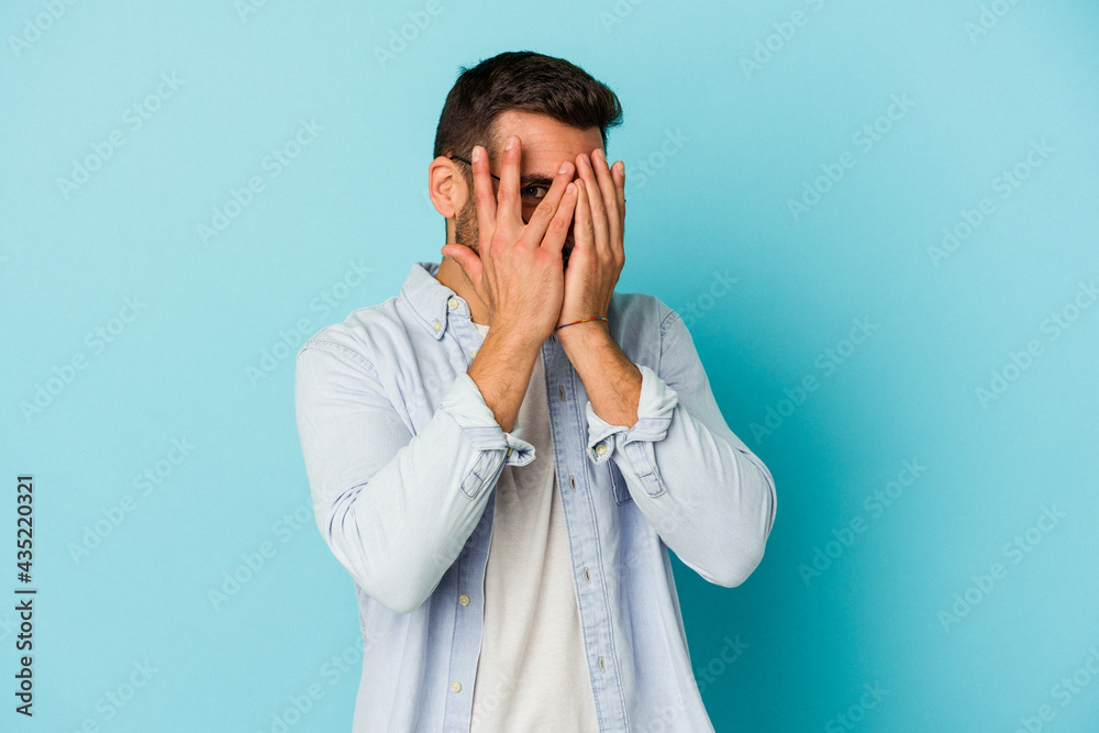 Young caucasian man isolated on blue background blink at the camera through fingers, embarrassed covering face. Young caucasian man isolated on blue background blink at the camera through fingers, embarrassed covering face.