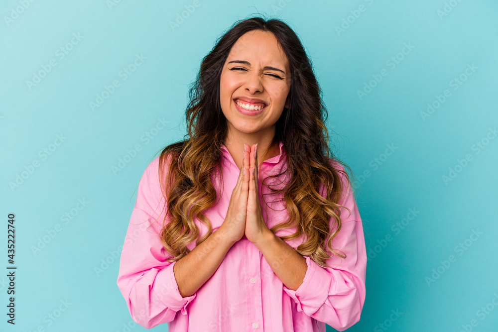Young mexican woman isolated on blue background holding hands in pray near mouth, feels confident.