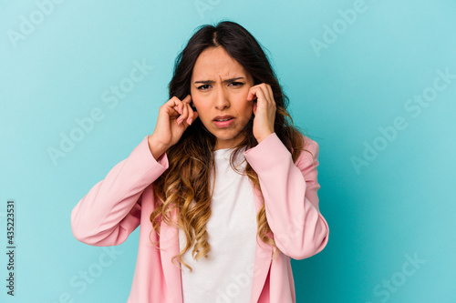 Young mexican woman isolated on blue background covering ears with fingers, stressed and desperate by a loudly ambient.