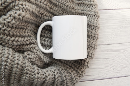 Cozy winter mood, white ceramic coffee mug mockup for design presentation, cup on grey warm scarf, white wooden table background.
