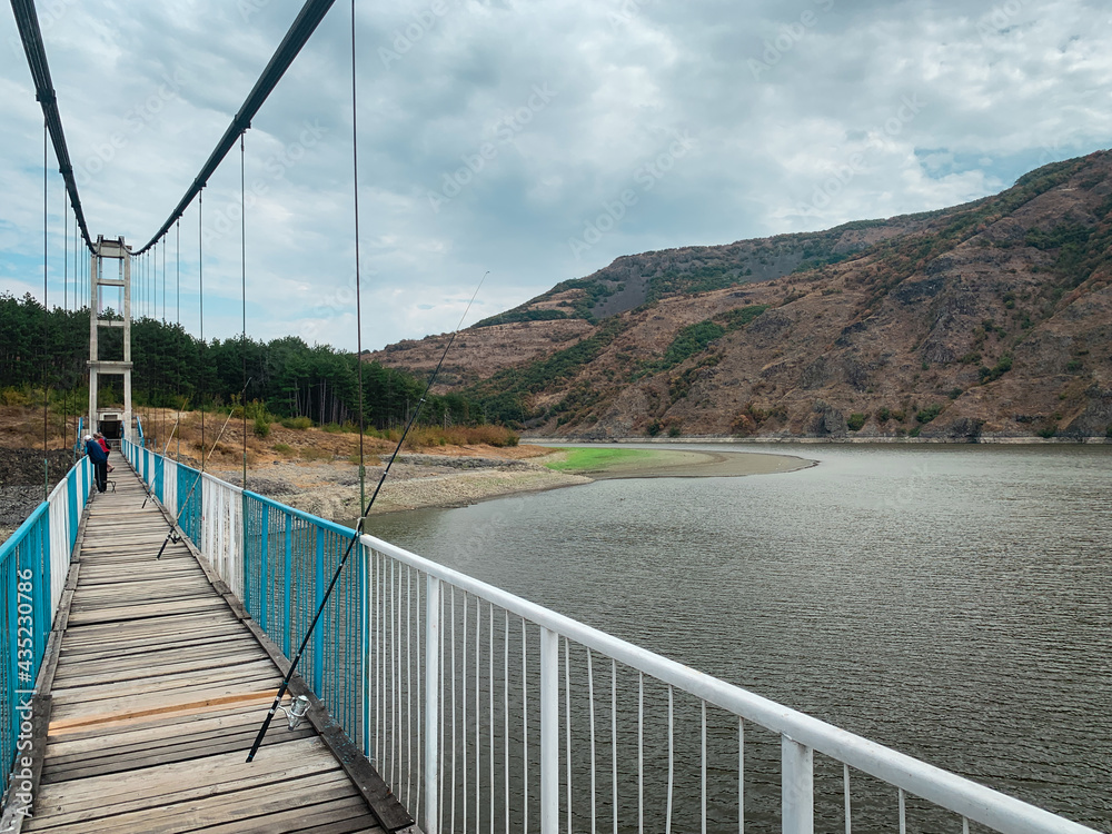 Fishing rods and fishermen standing on steel cable suspension bridge over big lake next to mountains