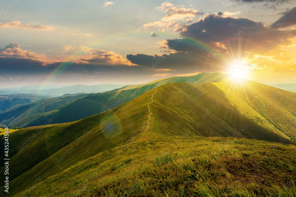mountain landscape in summer at sunset. grassy meadows on the hills rolling in to the distant peak beneath a rainbow in evening light