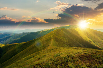 mountain landscape in summer at sunset. grassy meadows on the hills rolling in to the distant peak beneath a rainbow in evening light
