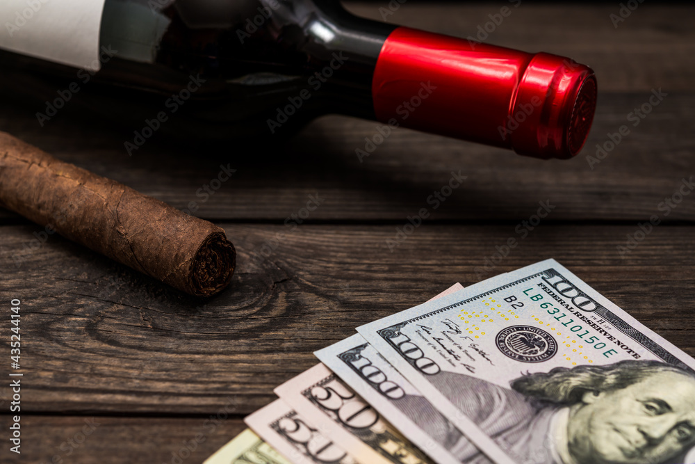 Bottle of red wine and cuban cigar with money on an old wooden table. Close up view, focus on the cuban cigar