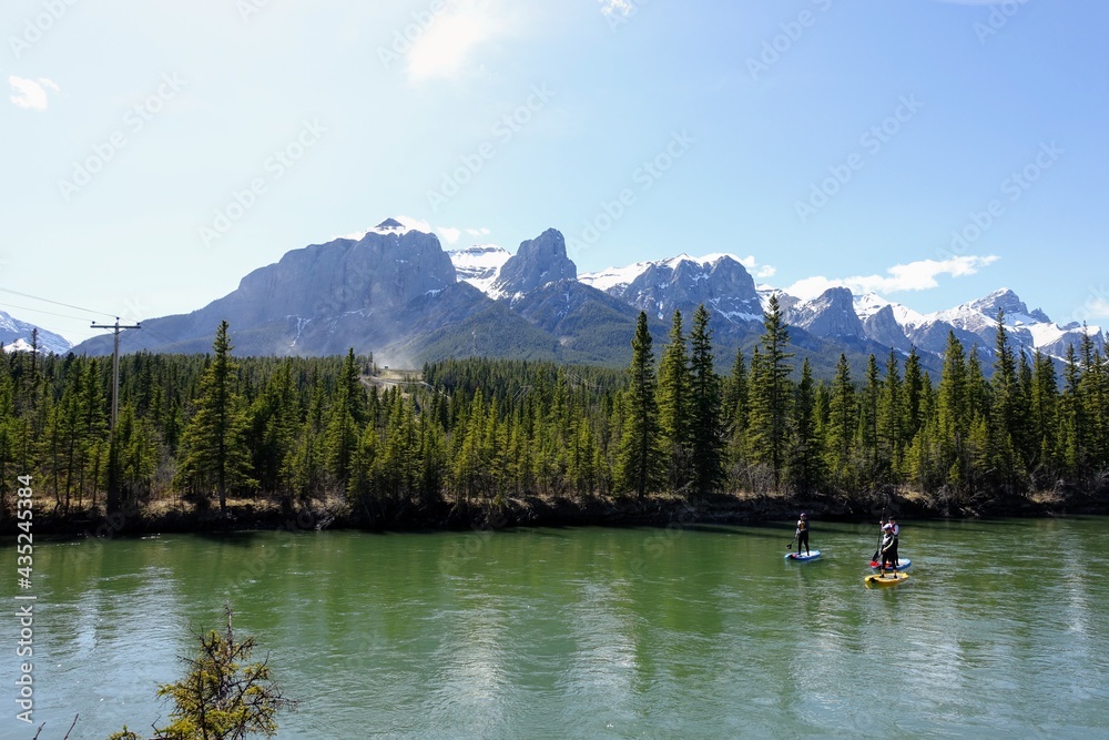 A group of people paddle boarding down the Bow River surrounded by forests and mountains in Canmore, Alberta, Canada