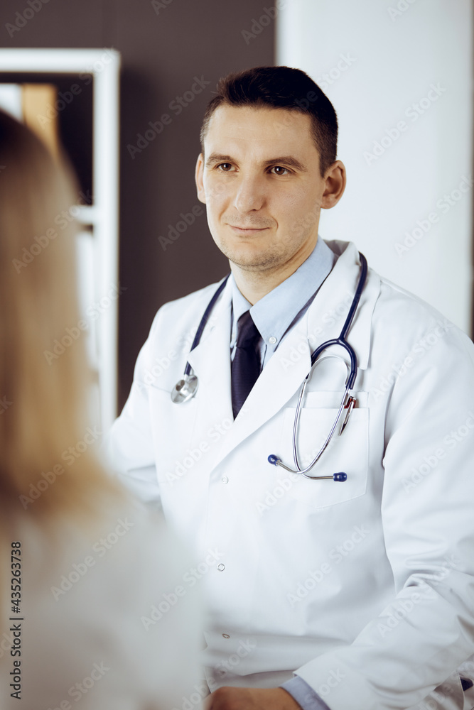 Friendly male doctor and patient woman discussing current health examination while sitting in sunny clinic. Perfect medical service in hospital Friendly male doctor and patient woman discussing current health examination while sitting in sunny clinic. Perfect medical service in hospital