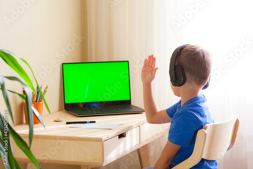 smart school boy in headphones sits at the table and looks at the computer. A child pulls a hand to answer. Online lesson at a distance learning. Green screen chroma key on the monitor.