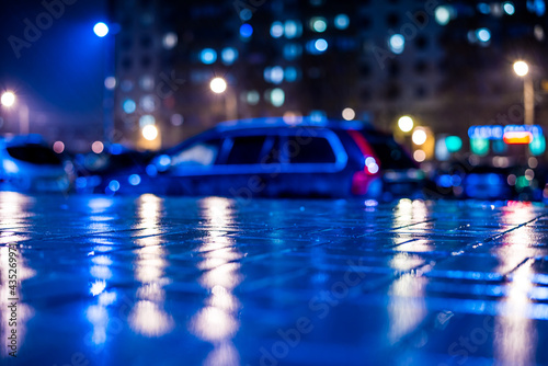 Rainy night in the big city, car parked next to the house, light from the the windows of the house is reflected in the asphalt. View from the sidewalk level paved with bricks © Georgii Shipin