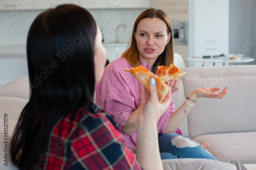Two young women talking and eating delicious pepperoni pizza while sitting at home. The concept of a carefree and happy life. High quality photo