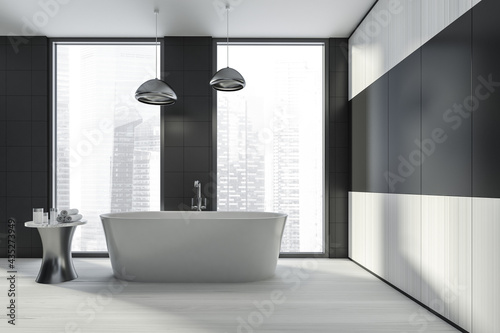Modern design bathroom interior with white oval bathtub  silver faucet  lamp. Small table with towel and cosmetics. Panoramic window with skyscrapers city view. Wood materials.