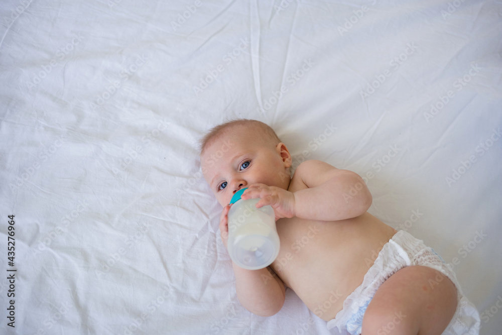 the baby boy is a blonde with blue eyes in a diaper lying on a white blanket on the bed and eating a mixture from a bottle. Artificial feeding the baby boy is a blonde with blue eyes in a diaper lying on a white blanket on the bed and eating a mixture from a bottle. Artificial feeding