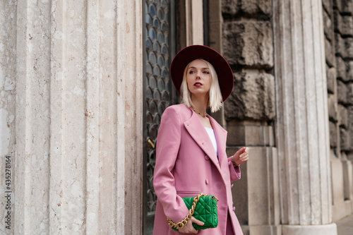 Fashionable woman wearing elegant pink blazer, holding green quilted leather bag with chunky chain, posing in street of European city. Copy, empty space for text