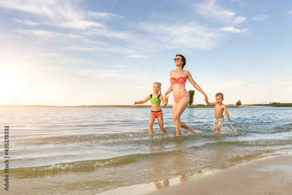 Naklejka premium Young adult attractive slim sporty mother enjoy having fun running water by lake or sea sand breach with two cute little siblings against blue sky on summer day. Summertime family vacation concept