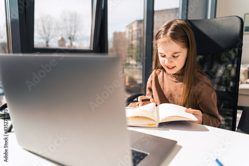 Cute little schoolgirl studies at home near laptop