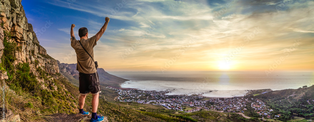 Fototapeta premium Victory and success after a hike up Table Mountain at sunset. Looking over the bay with a vibrant orange sky - Great outdoors adventure and travel holiday destination, Cape Town, South Africa