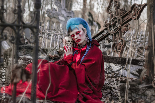 Sensual gothic beauty in the cemetery. Beautiful model with a skull sitting at the cross. Unusual red makeup and cape.