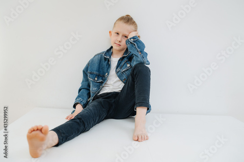 Portrait of a reluctant nine year old boy in a jeans suit. He has blond hair in a bun. Sitting on a table. Over white. photo