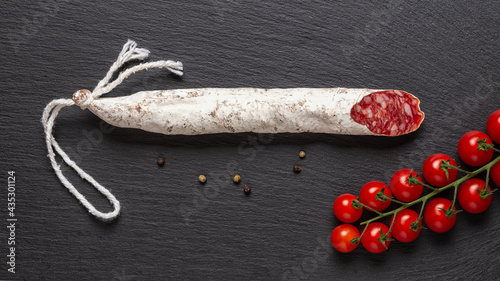 Thin catalan fuet sausage and bunch of red cherry tomatoes on a black slate surface. Wide image of traditional spanish dry-cured pork salami with white noble mold. Tasty meat delicacy. photo