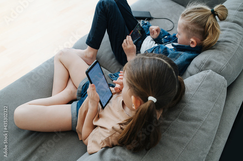 Pair of little siblings sitting next to each other. They are spending their free time watching their phones on a couch, playing or watching cartoons. In the living room.