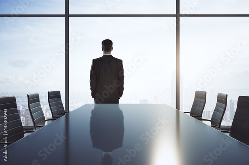 Empty meeting room with big conference table, chairs around and businessman back looking through big window on bright sky. photo
