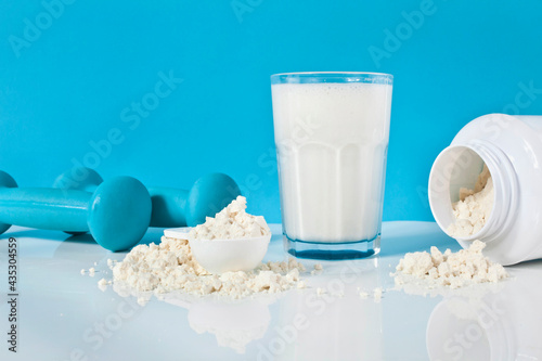 Glass of protein cocktail and protein powder in the spoon and container with blue dumbbells on a blue background.