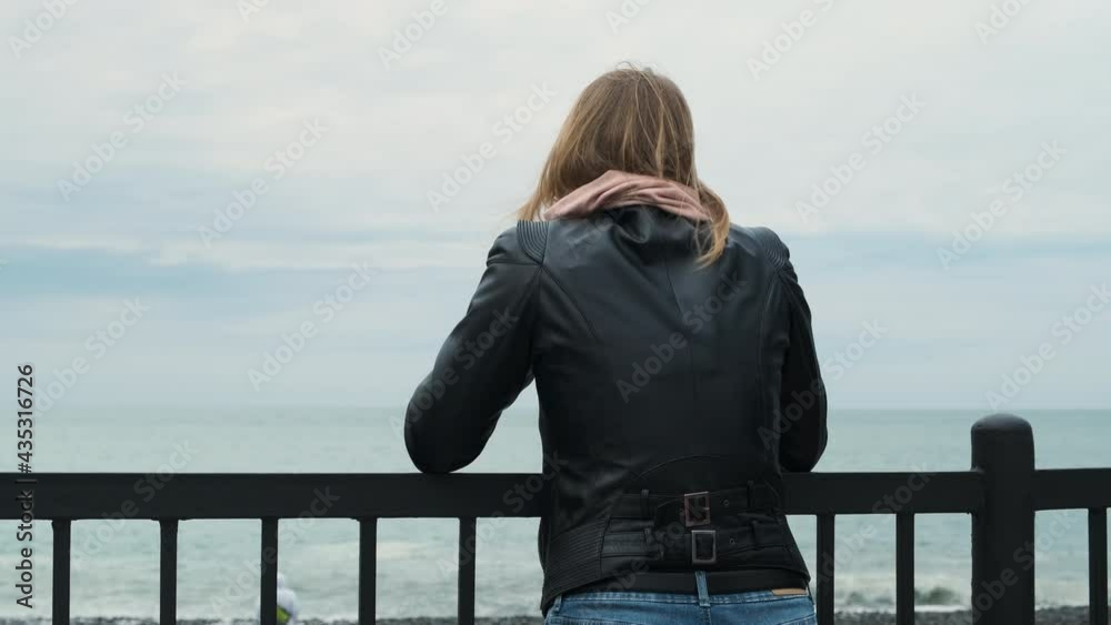 Rear view of white-haired blonde woman in black leather jacket leaning on railing against backdrop of sea or ocean and looking into distance or rummaging through phone and looking for information. Rear view of white-haired blonde woman in black leather jacket leaning on railing against backdrop of sea or ocean and looking into distance or rummaging through phone and looking for information.