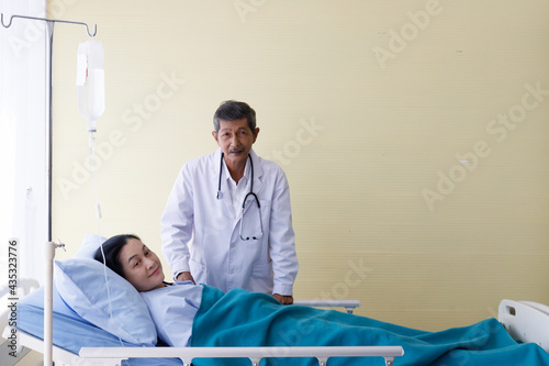Asian male elderly gray hair and mustache doctor in white lab coat uniform with stethoscope visit female patient lay down smile on bed cover by blanket inject with normal saline solution drip in ward