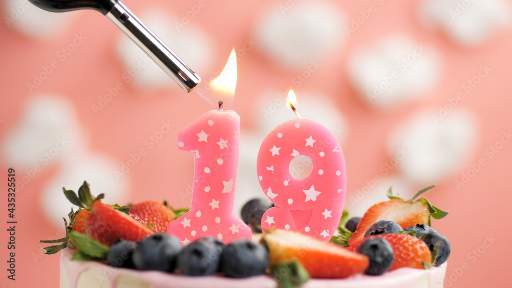 Birthday cake number 19, pink candle on beautiful cake with berries and lighter with fire against background of white clouds and pink sky. Close-up