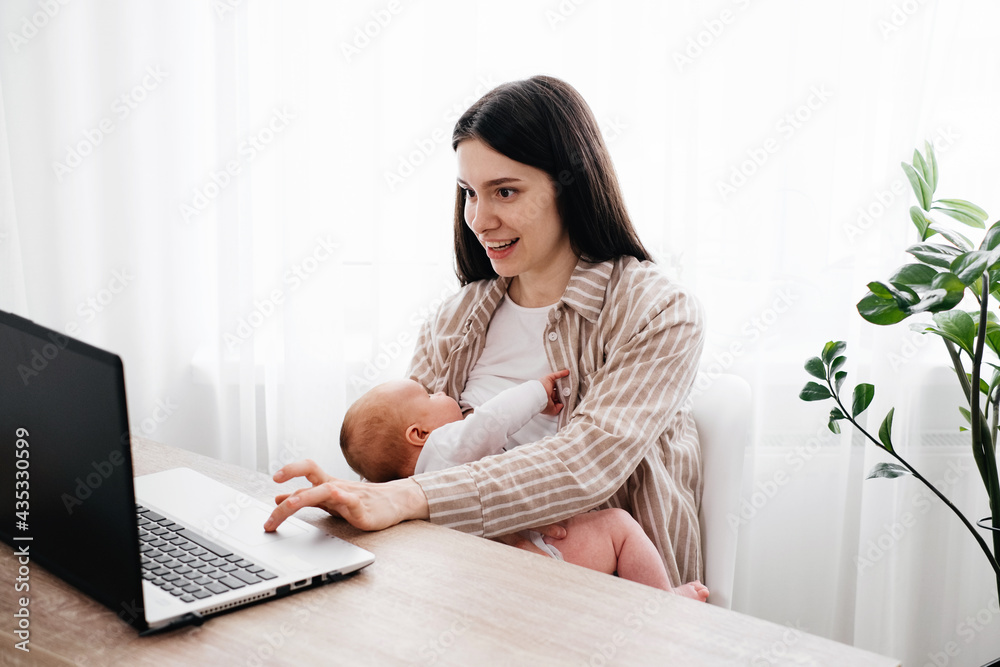 Working and breast feeding woman with infant on her hands near laptop at home over the window. Studying on-line mother near computer with baby boy. Working from home mom with child Working and breast feeding woman with infant on her hands near laptop at home over the window. Studying on-line mother near computer with baby boy. Working from home mom with child