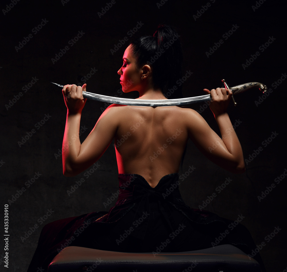 Young brunette woman warrior, martial arts fighter sits in skirt and topless back to camera holding sword on shoulders, looking aside over dark background. Martial arts and beautiful women concept Young brunette woman warrior, martial arts fighter sits in skirt and topless back to camera holding sword on shoulders, looking aside over dark background. Martial arts and beautiful women concept