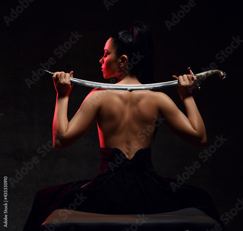 Young brunette woman warrior, martial arts fighter sits in skirt and topless back to camera holding sword on shoulders, looking aside over dark background. Martial arts and beautiful women concept