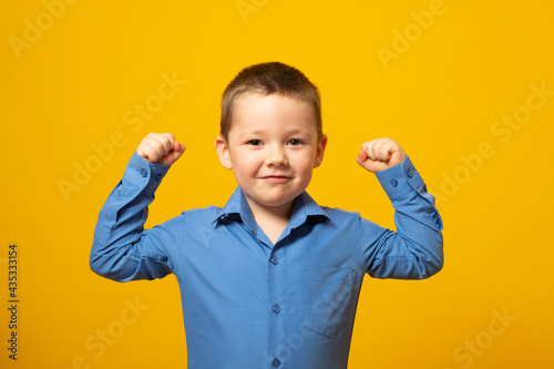 Cute little boy showing his hand biceps muscles strength isolated on yellow background.