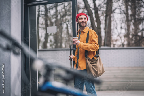 Man in a red hat and orange jacket with a coffee cup in hands looking cheerful