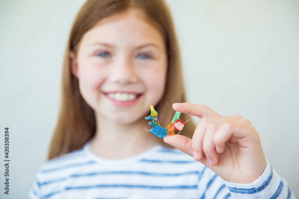 Smiling little child girl holds metal bright and colorful plate for align the teeth and bite. Orthodontist treats teeth - obrazy, fototapety, plakaty 