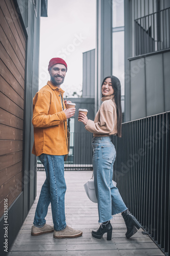 Young man and woman in casual clothes looking positive while having coffee outside