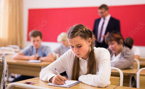 Portrait of focused teenage schoolgirl writing lectures in workbooks in classroom during lesson