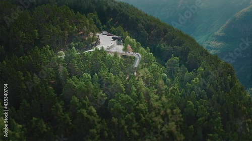 A narrow asphalt road winding uphill through the dense forest, leading to the famous Stegastein viewpoint. The viewpoint platform hanging from the cliff overlooking the fjord and the vast valley. photo