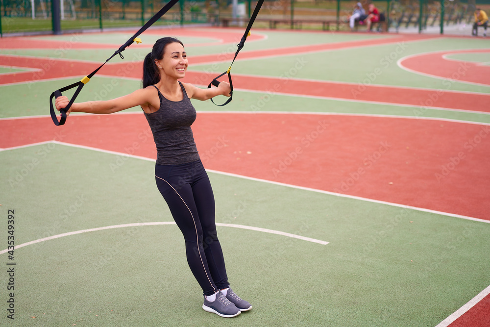 Girl athlete training using on sport ground. Mixed race young adult woman do workout with suspension system. Healthy lifestyle. Stretching outdoors playground.