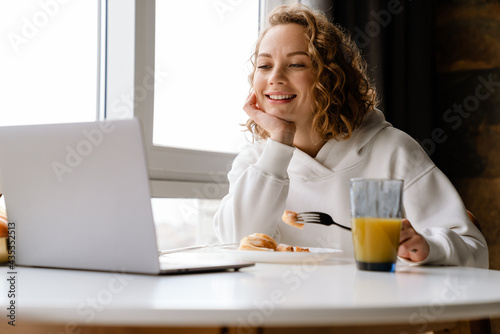 Young white woman using laptop while having breakfast at home