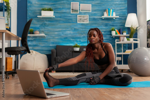 Sporty fit black woman doing stretching flexibility exercise reaching toes sitting on fitness mat in home living room watching tutorial video on laptop screen. Workout and healthy lifestyle.