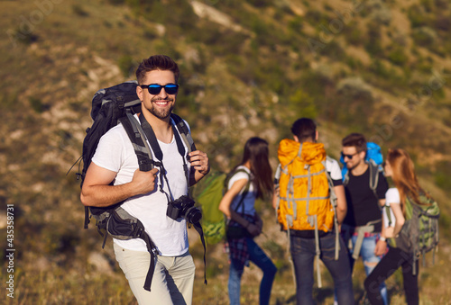 Handsome smiling male hiker with backpack and camera standing in nature against the background of his tourist friends. Concept of hiking and active lifestyle. Blurred background.