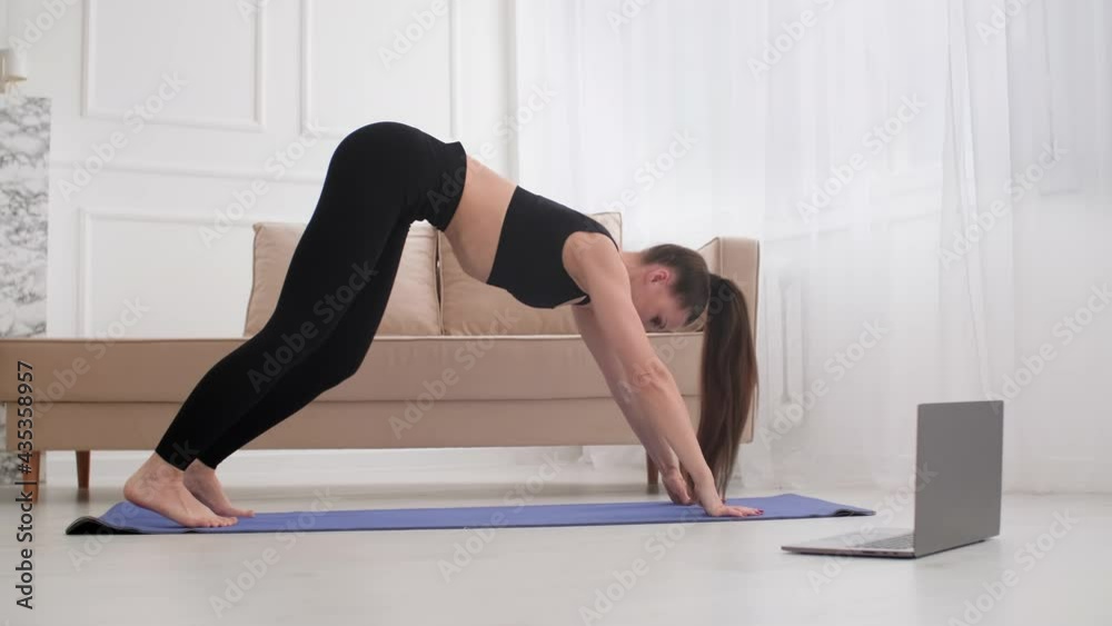 Woman practicing yoga exercise in living room with laptop. Young female doing stretching exercise on yoga mat. Brunette woman doing sports at home. 4K, UHD Woman practicing yoga exercise in living room with laptop. Young female doing stretching exercise on yoga mat. Brunette woman doing sports at home. 4K, UHD