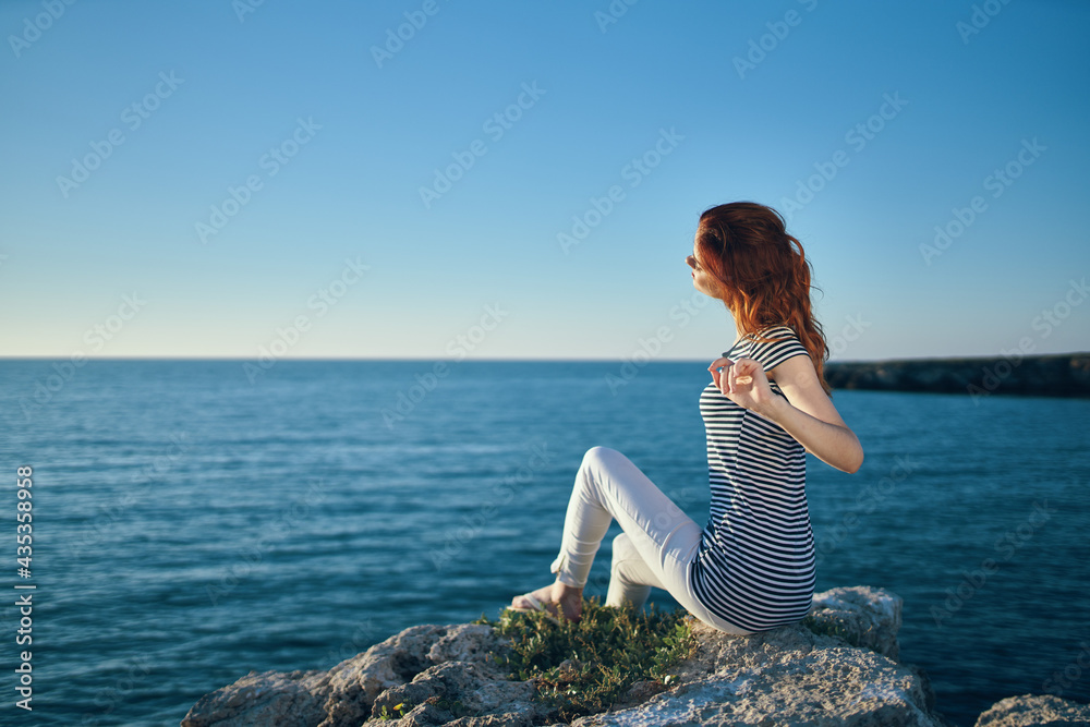 happy woman in t-shirt trousers sits on a large stone near the sea in summer