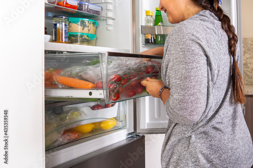 Young woman searching in a fridge draw for vegetables for cooking a salad. Open refrigerator drawer with tomatoes and carrots, food leftover containers on the shelves. Real vegetarian household fridge