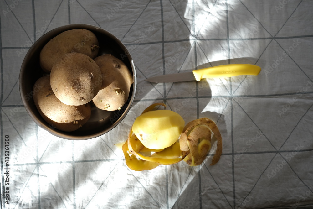 Potatoes in bowl, peeled potatoes, potato peels and knife on fabric.