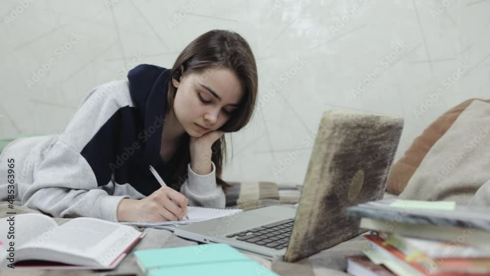 Thoughtful woman taking notes and studying online at home. Pensive young female student lying on bed and writing in notebook during remote lesson via laptop Thoughtful woman taking notes and studying online at home. Pensive young female student lying on bed and writing in notebook during remote lesson via laptop