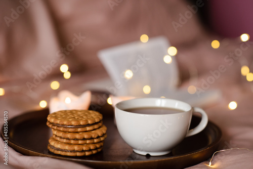 Cup of fresh black tea with stack of biscuits with chocolate filling on tray with burning candles and open paper book in bed over glow lights. Cozy home atmosphere.