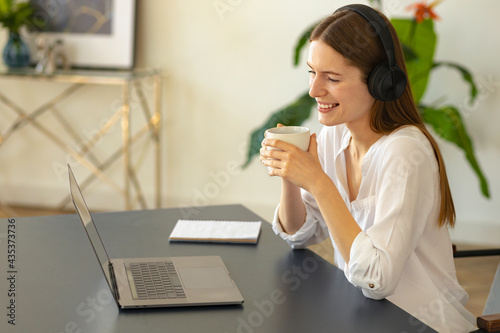 Young caucasian woman in headphones using laptop  looking at screen  happy woman with cup coffee watching funny video on the internet during lunch break